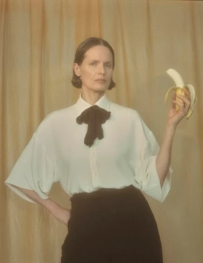 Woman in white shirt with black bow tie holding a peeled banana against a draped yellow background