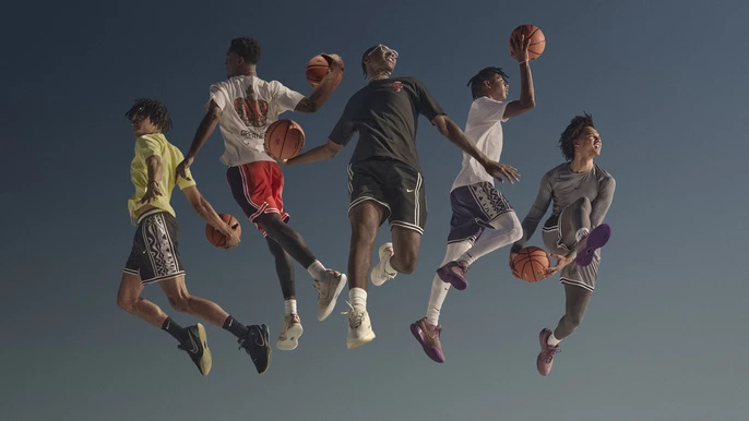 Five adult males leaping in the air playing with basketballs against a plain dark blue background, wearing athletic attire and sneakers.