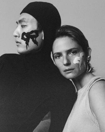 Monochrome portrait of an East Asian man in a black hood and a woman in a pale top with painted facial markings.