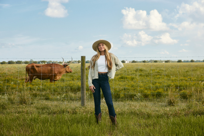 Adult female in a hat poses in a rural field with a brown longhorn cow behind a fence under a blue sky with scattered clouds.