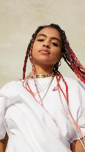Portrait of a young adult female with braids and jewelry, wearing a white t-shirt, against a neutral beige background in eye-level view.