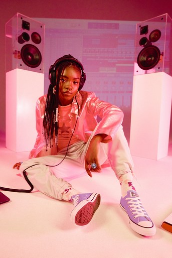 Young adult woman with headphones sitting in pink-lit studio between speakers, wearing pink jacket and sneakers, looking at camera.