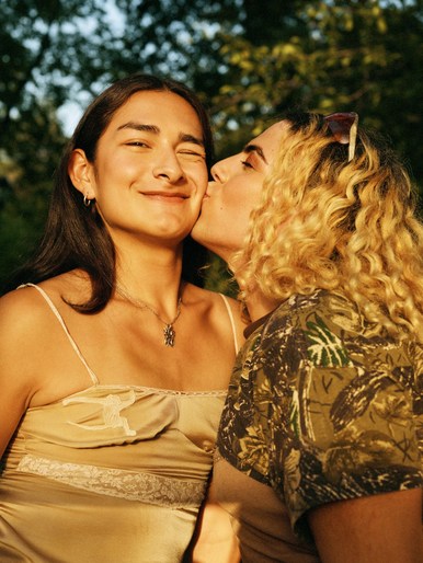 Two young women in outdoor setting, one kissing the other's cheek in warm light. The woman being kissed is smiling in the golden hour.