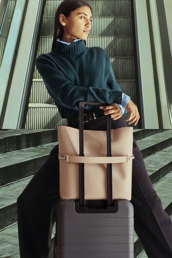 Brunette woman poses with luggage and a tote bag in front of an escalator in an indoor setting with soft, diffused lighting.