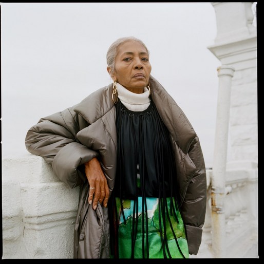 Mature woman in a puffer coat stands outdoors against a cloudy sky. She wears a turtleneck, layered top, and gold earrings.