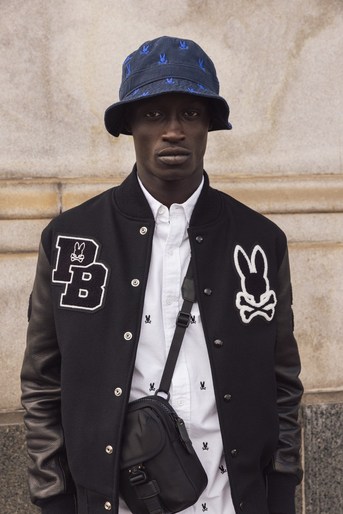 Man in stylish bomber jacket and bucket hat with logo stands against neutral wall, showcasing fashion and urban style in portrait shot.