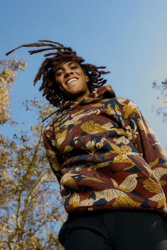 Smiling Black male in a floral hoodie looking up at the camera in a casual outdoor setting with trees and a blue sky backdrop.