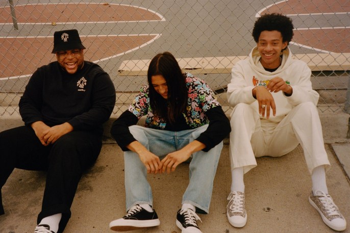 Three smiling friends sit together on an outdoor basketball court wearing casual streetwear fashion with natural lighting and a chain link fence.