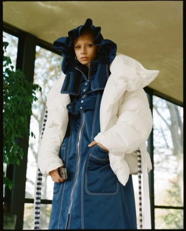 Editorial fashion photograph of a young adult woman wearing a navy blue dress with white puffer sleeves and a large ruffle hood indoors.