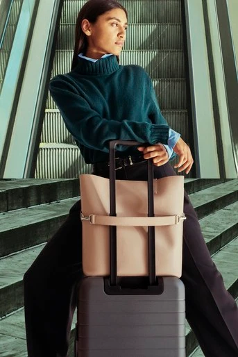 Woman in dark green turtleneck leaning on a pink tote bag resting atop a grey suitcase outdoors near steps