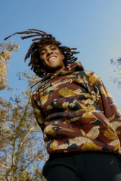 A smiling man with dreadlocks wearing a floral hoodie shot from a low angle against a clear blue sky.