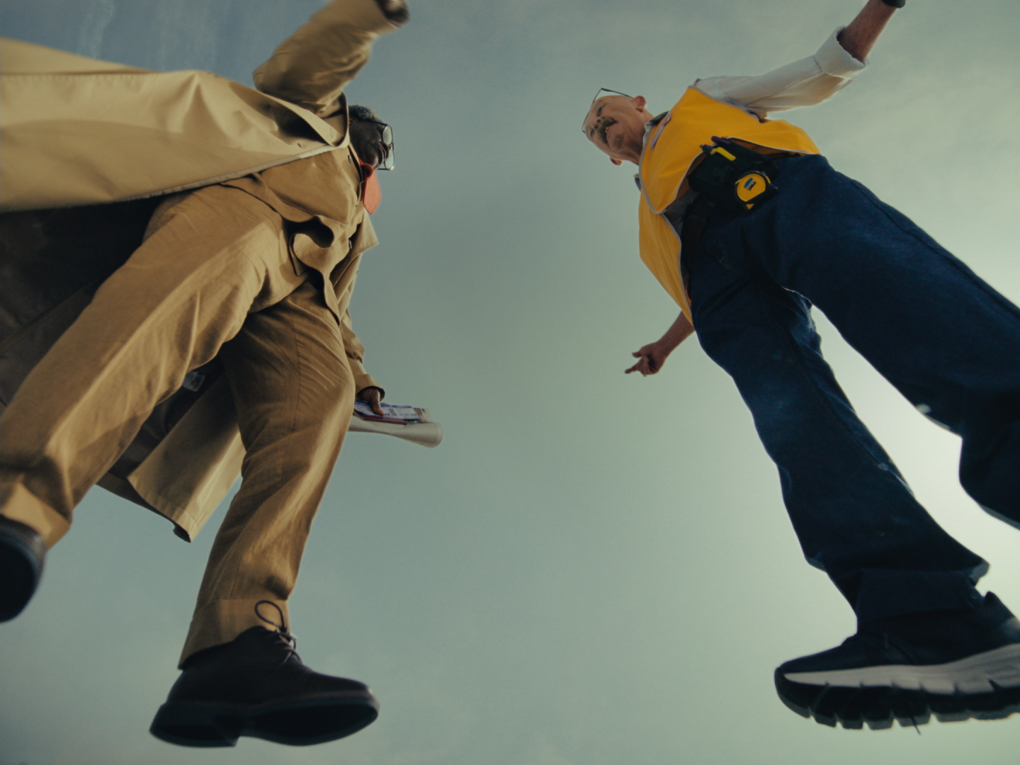 Two men in a low angle shot against a light blue sky. One in a trench coat and the other in a safety vest looking downwards.