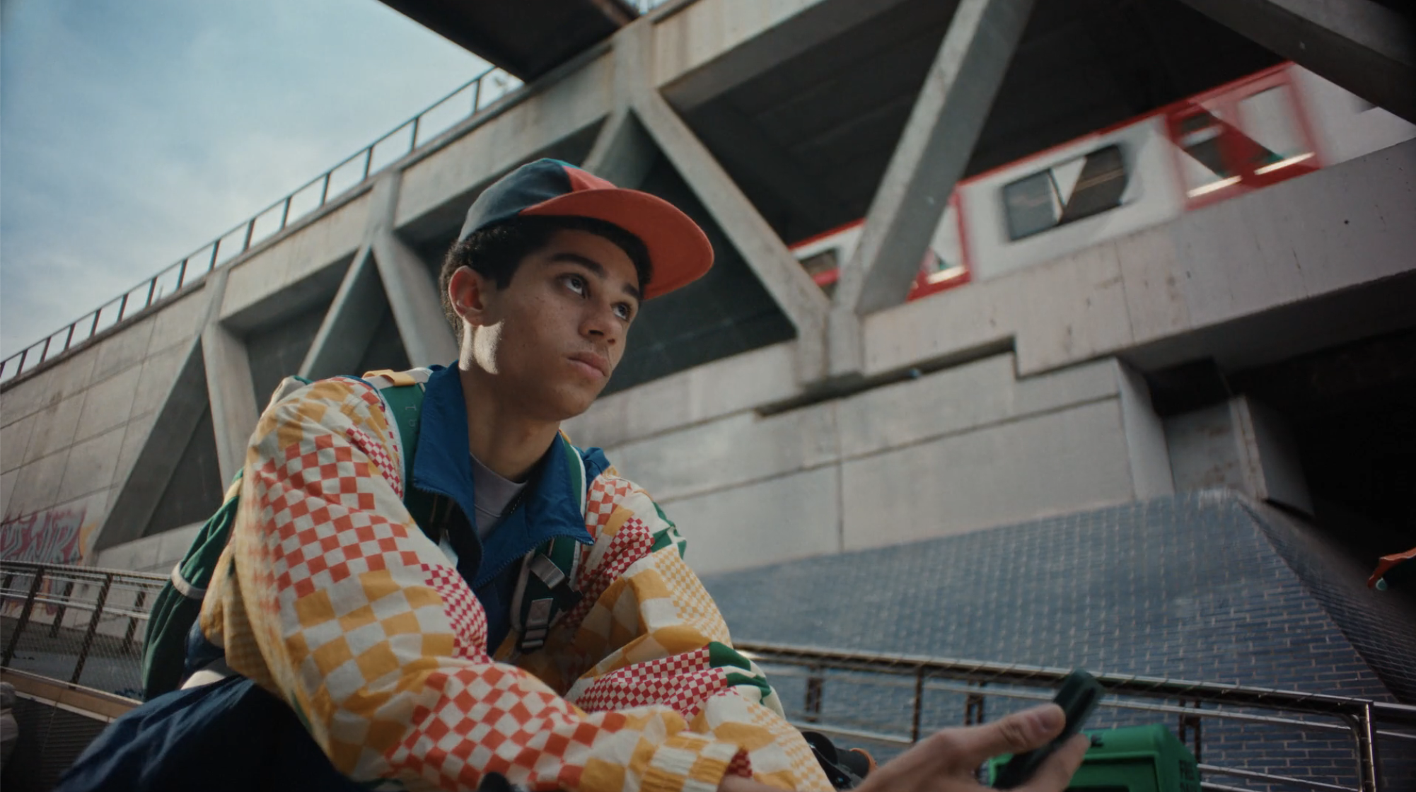 Young adult wearing a patterned jacket sits outdoors near a subway line with an electronic device in hand on an overcast day.