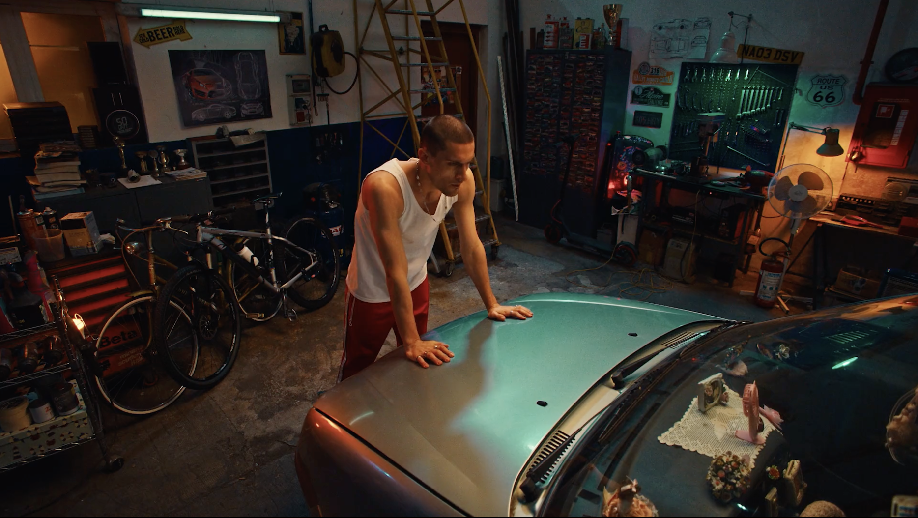 Man in white tank top leans on hood of car in cluttered garage with tools, bikes, posters, and muted lighting, creating an edgy vibe.