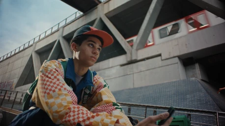 A young man in a checkered jacket and cap seated outdoors near an elevated concrete structure as a train passes above.