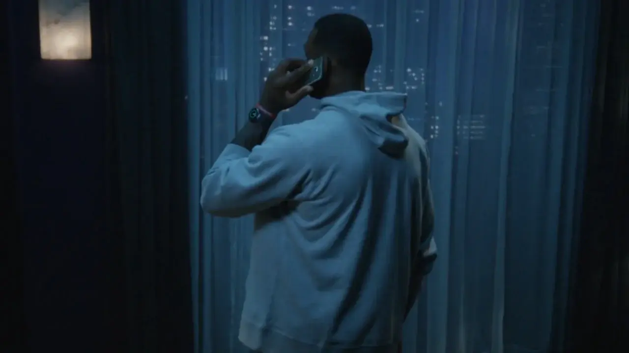 Man in grey hoodie talks on smartphone in dim indoor setting, city lights visible through sheer curtains in the background at night.