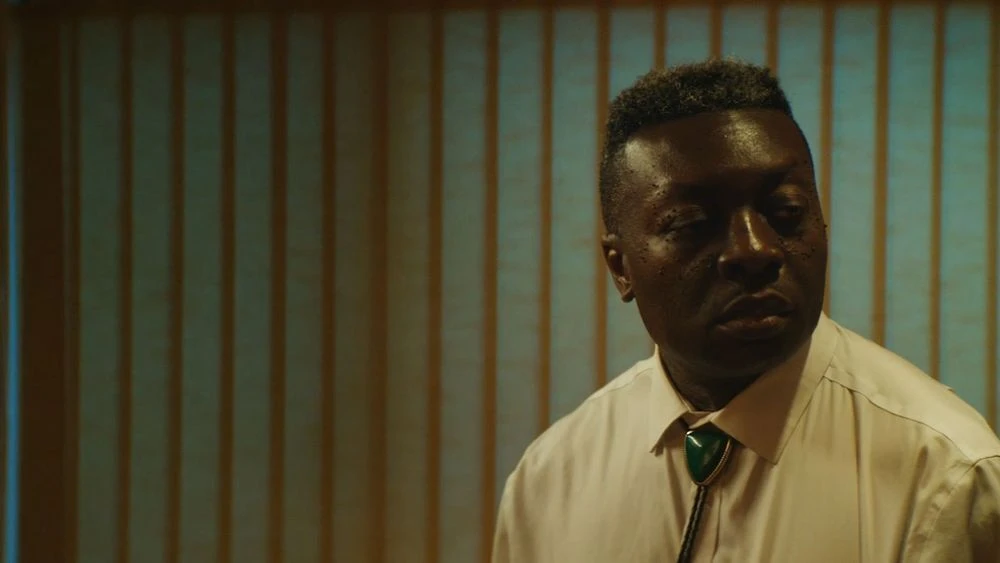 Portrait of an African American man in a white dress shirt and bolo tie against a vertically striped wall in a dimly lit setting.