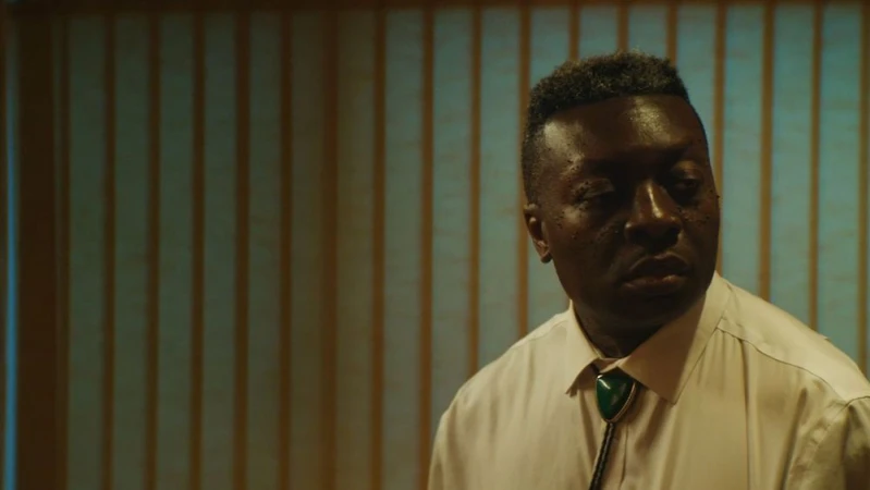 Portrait of an African American man in a white dress shirt and bolo tie against a vertically striped wall in a dimly lit setting.