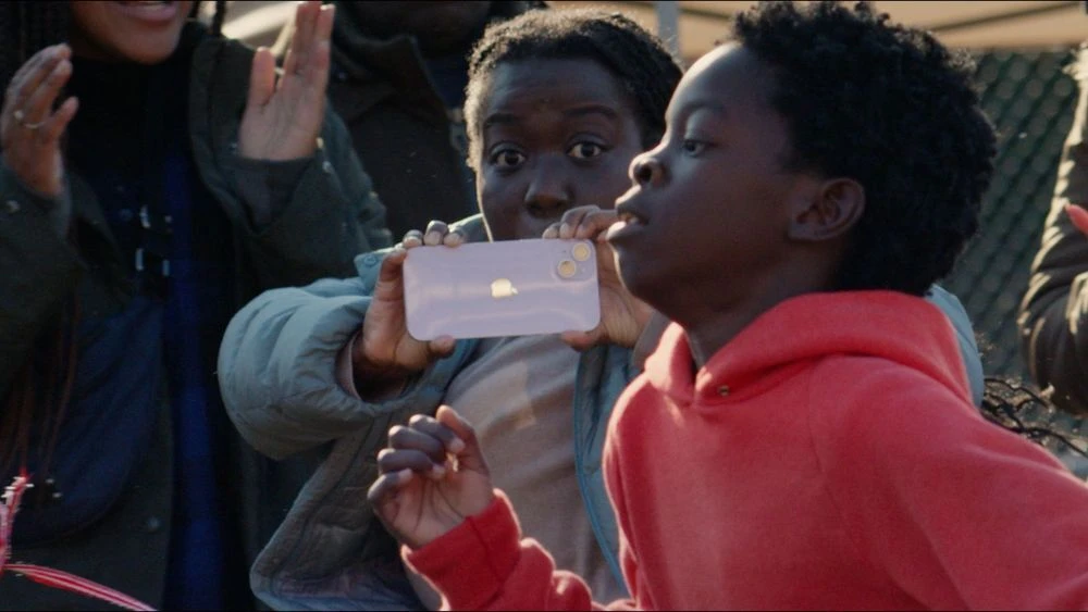 Two African-American children watching with phone. Child with orange hoodie smiling with focus. Casual outdoor scene with excitement.