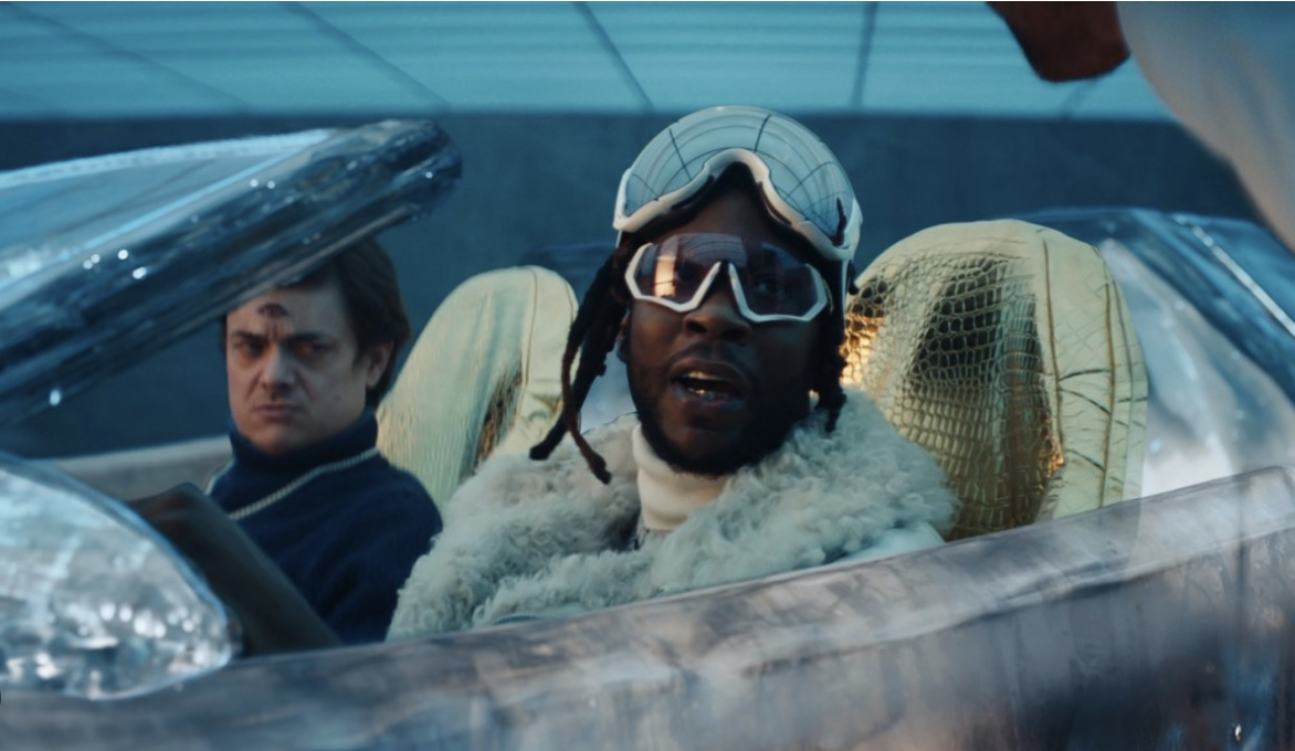 Two men inside an elaborate car with an unusual design; one man driving, the other looks at the camera wearing goggles and a fur coat.