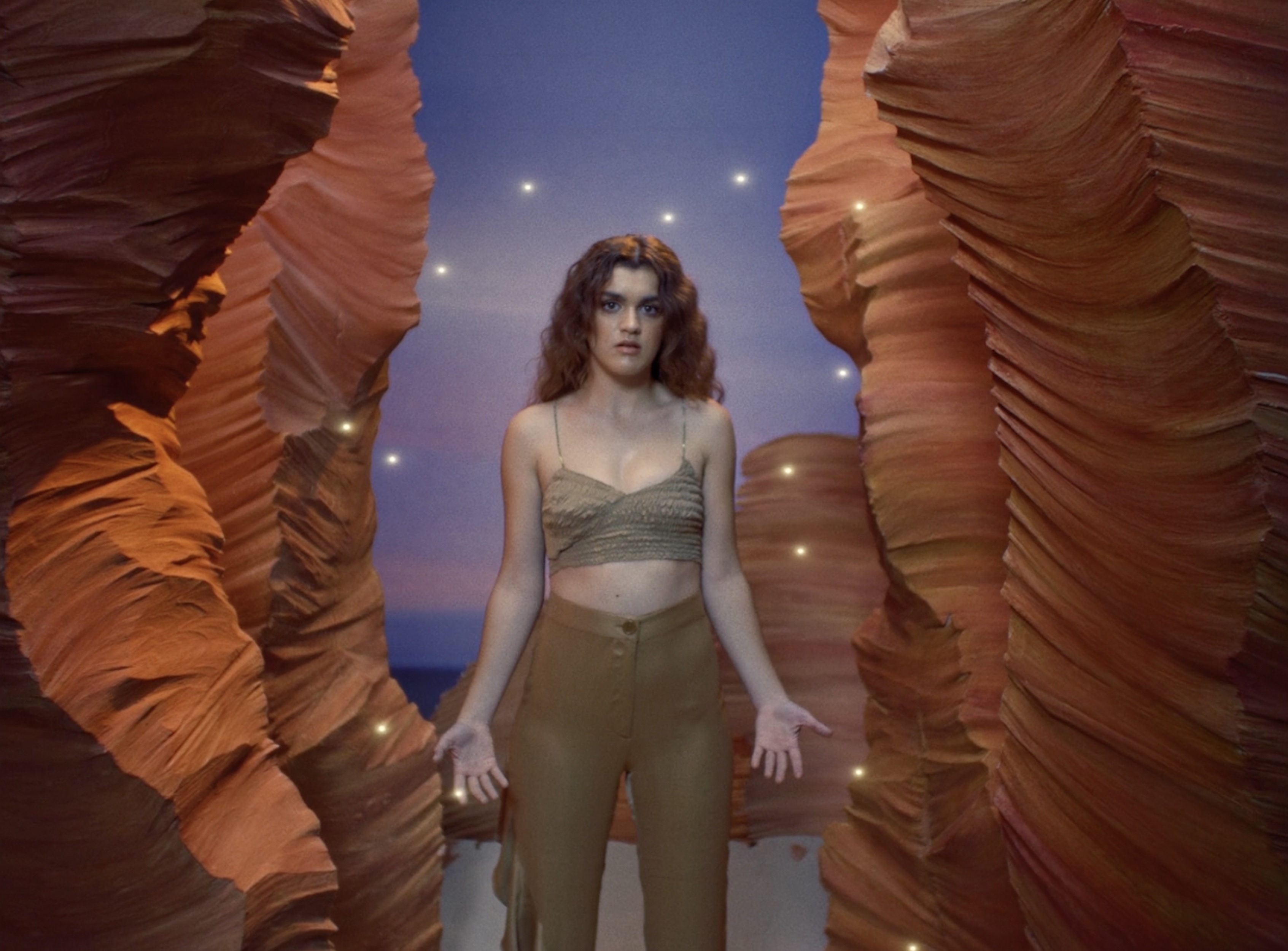 An adult woman stands in a desert canyon setting with brown hair, wearing a tan knit top and pants, posing with arms slightly outstretched.