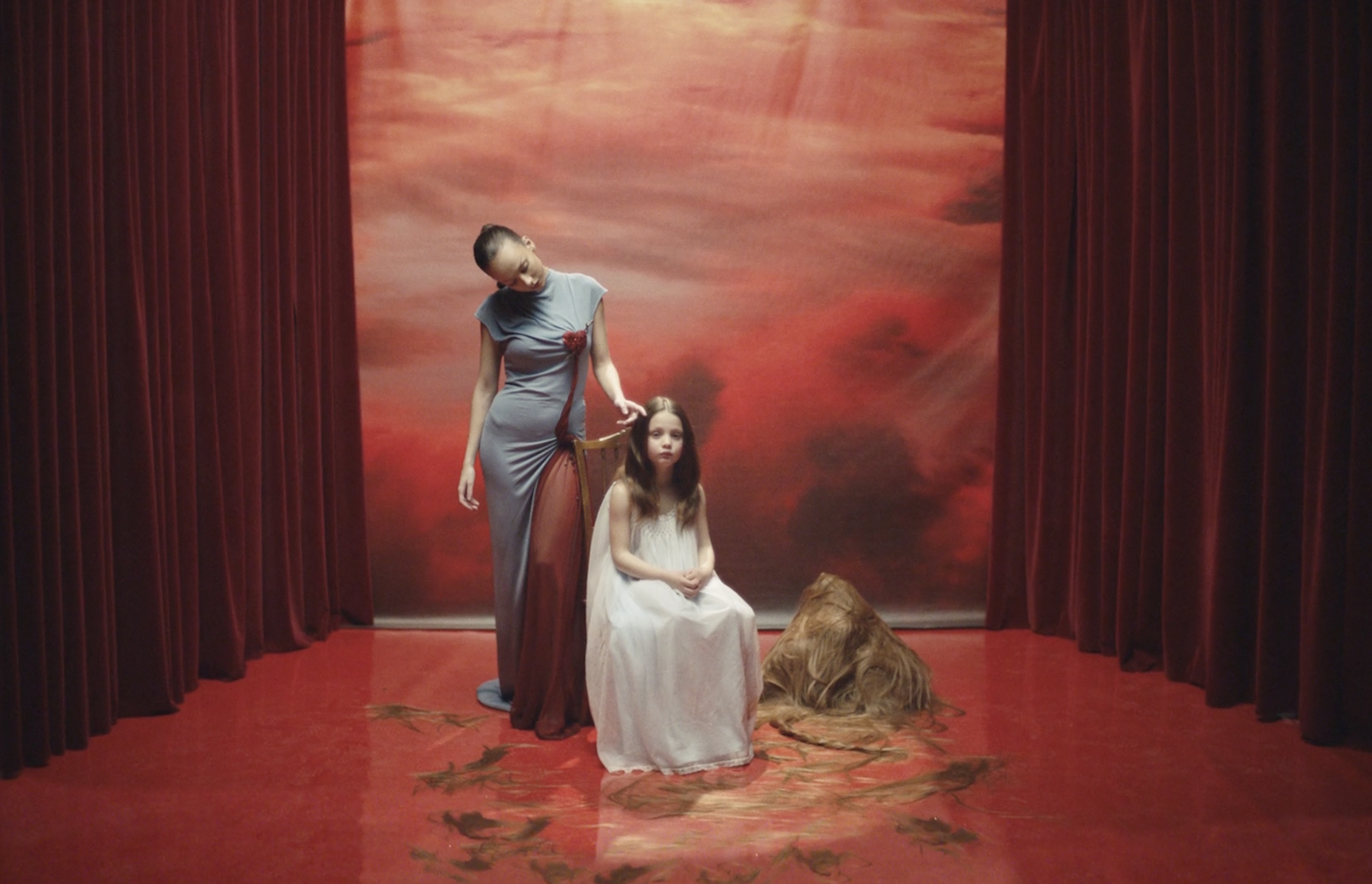 A woman in a grey gown stands beside a child in a white dress, both in a surreal scene with red curtains and a red cloudy backdrop.