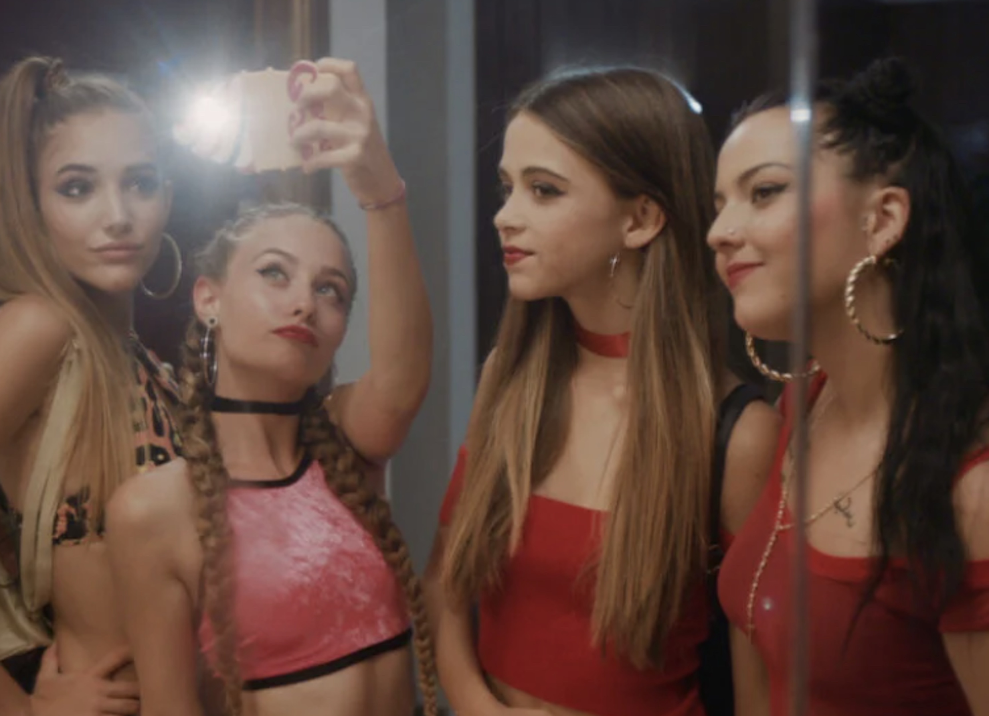 Five young women pose for a selfie, smiling and looking at the camera. They are indoors, with artificial lighting.