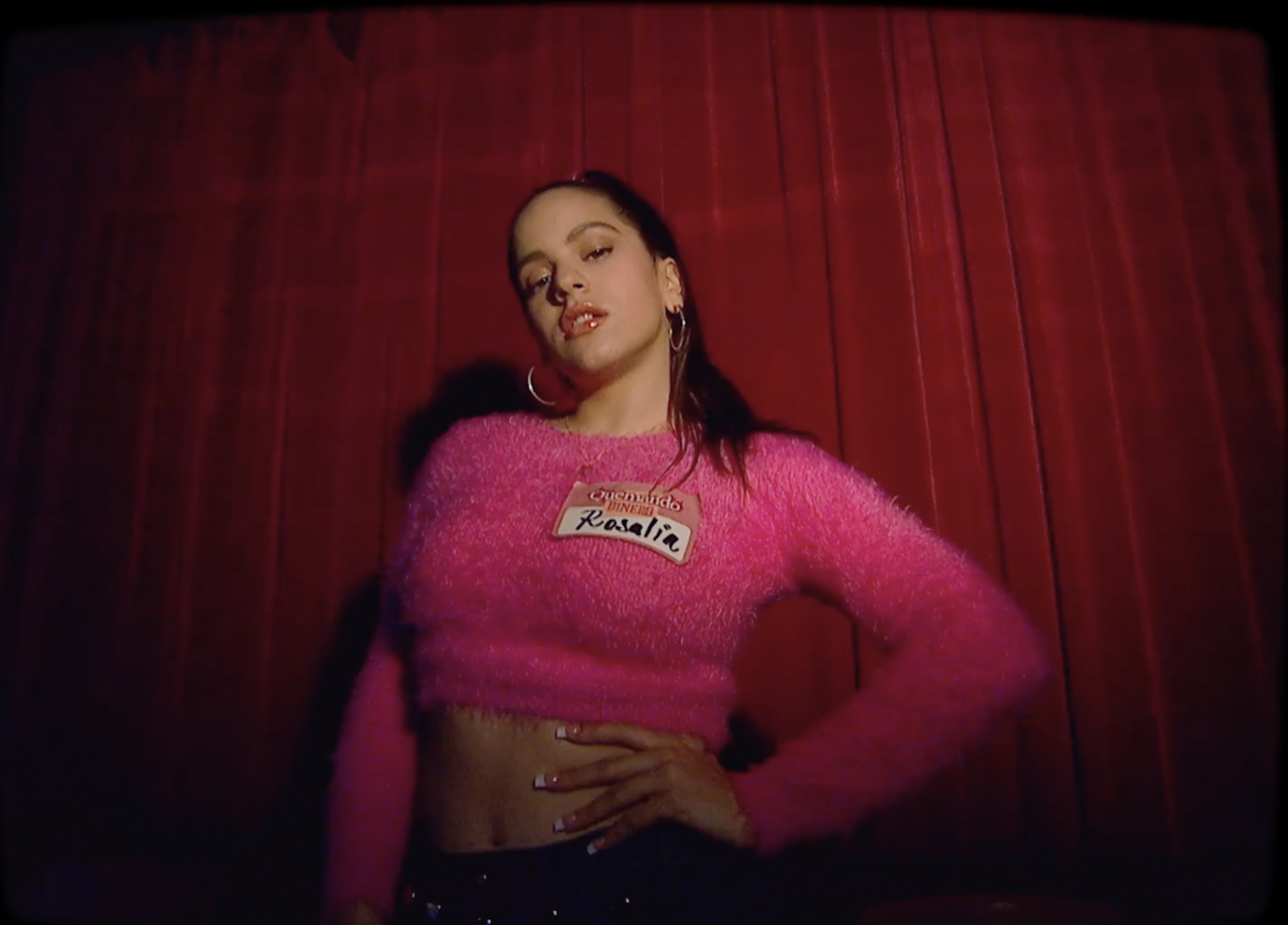 Rosalia poses in a pink top against a red curtain backdrop, showcasing a confident gaze and distinct fashion style.