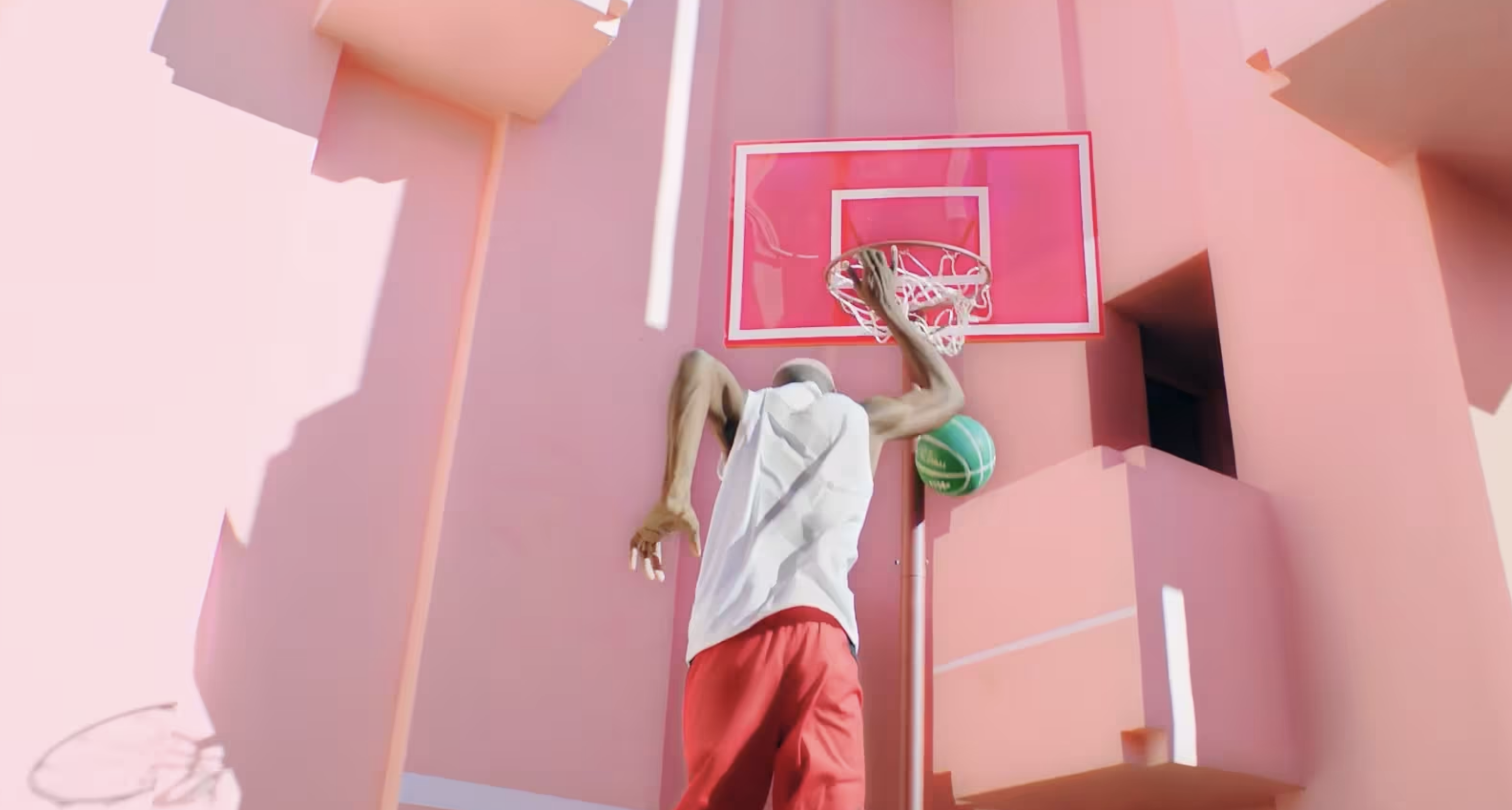 An athlete performs a basketball dunk near a coral-colored wall with contrasting shadows, showcasing athleticism and urban sport.
