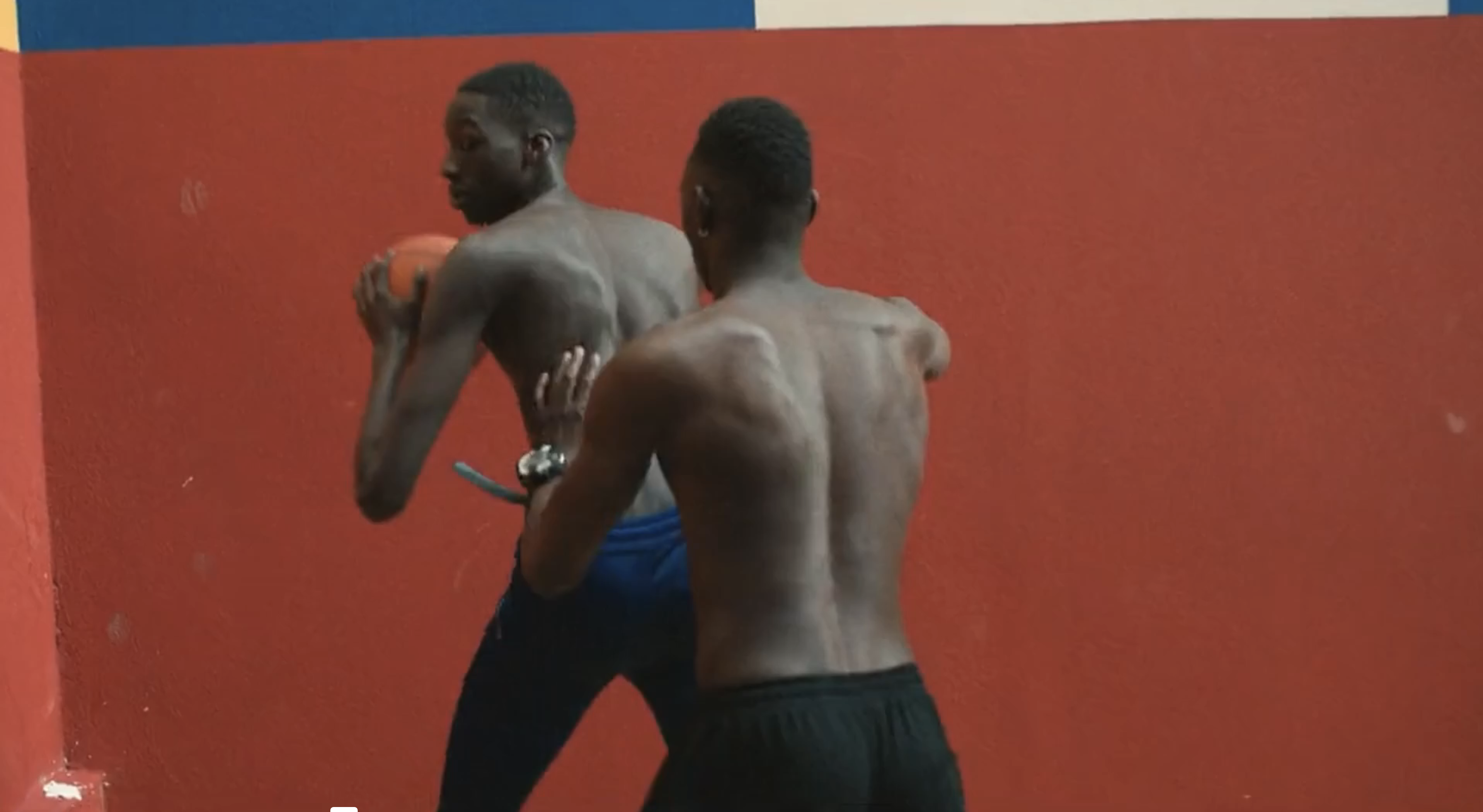 Two shirtless African-American men in athletic wear indoors, one holding basketball, in front of a red and blue wall. Gym setting.