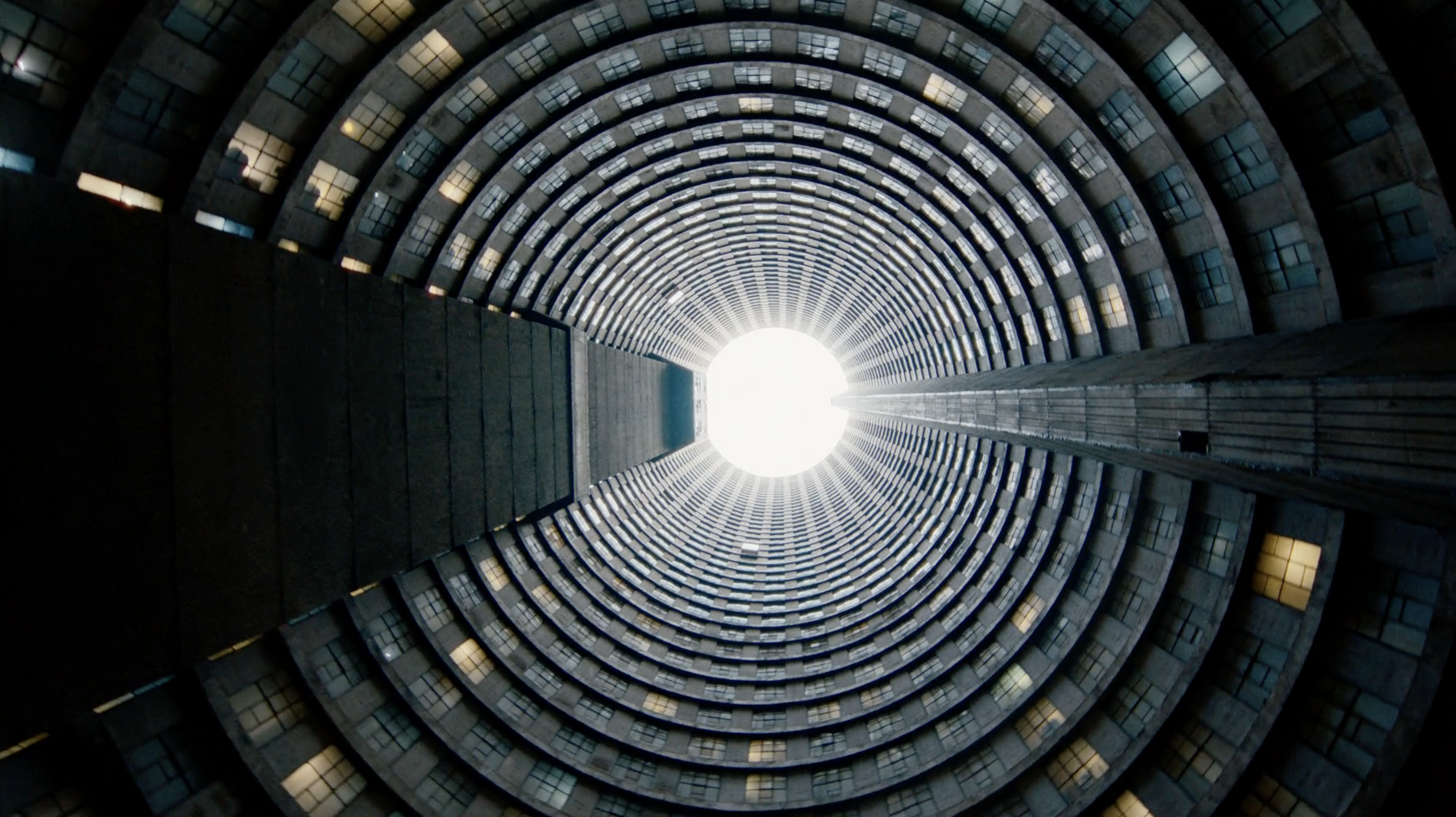 A high-angle shot of a cylindrical concrete building with many windows curving upwards to a bright, circular opening at the top.