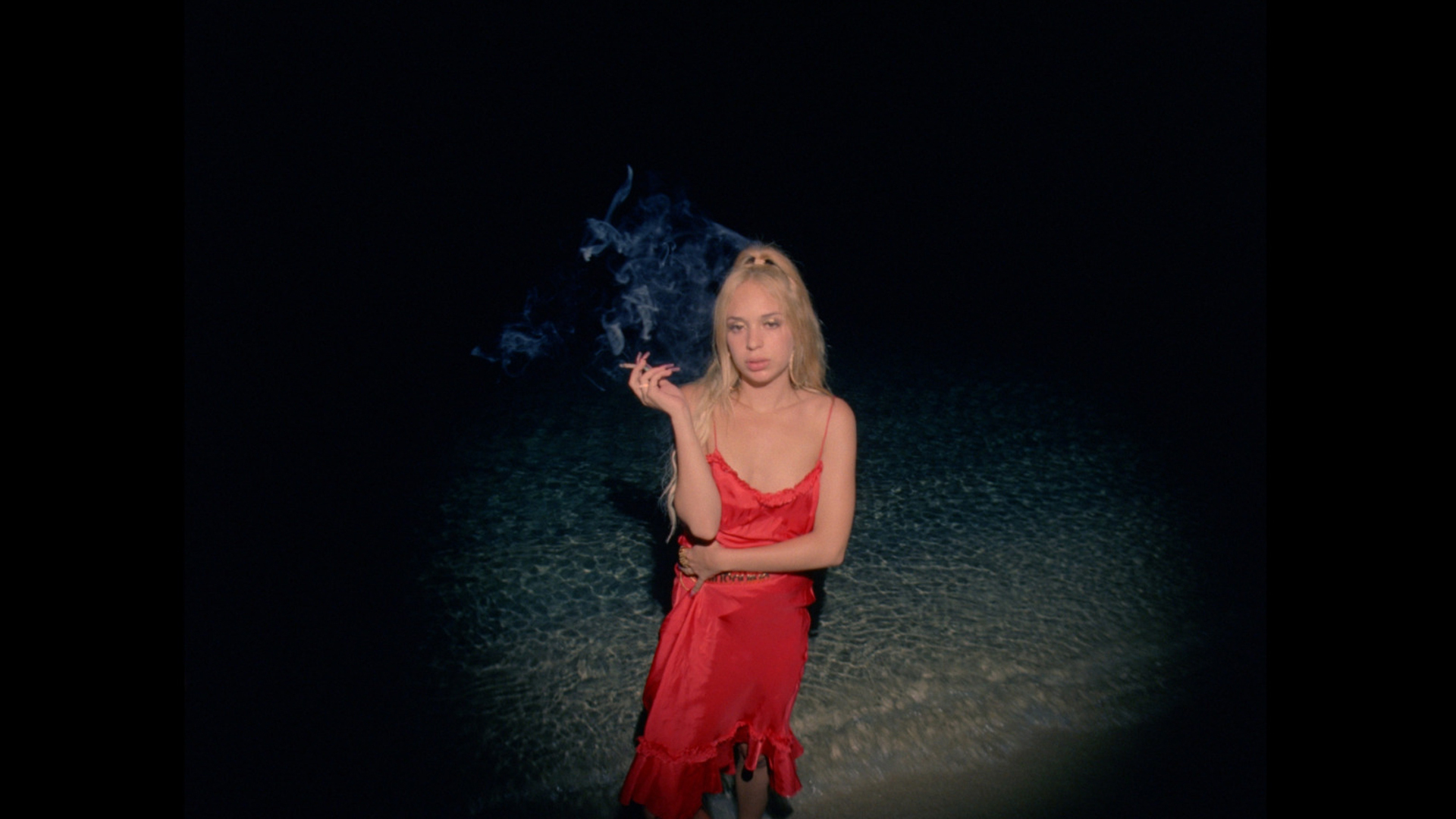 A blonde woman in a red dress stands in shallow water at night, smoking a cigarette, with smoke swirling in the dark background.
