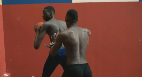 Two shirtless young black men playing basketball indoors against a red wall by Cyprien Clément-delmas
