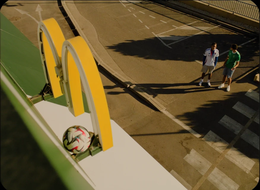 Two soccer fans wearing jerseys stand on a city street near a soccer ball and yellow arches, bathed in sunlight. Urban setting.