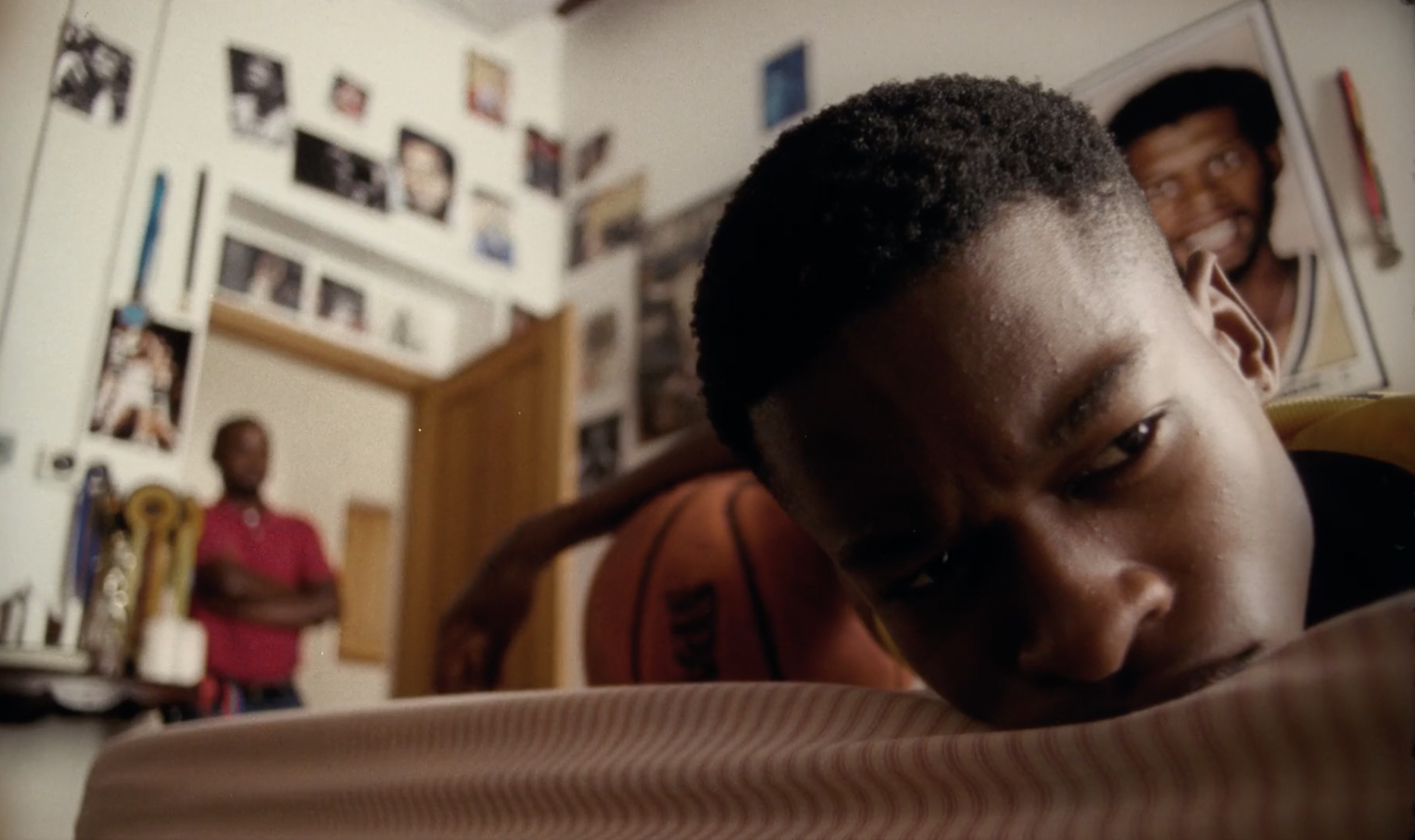 Close up of a teenage African American boy lying on a bed with a basketball, while an adult stands in a doorway in the background.