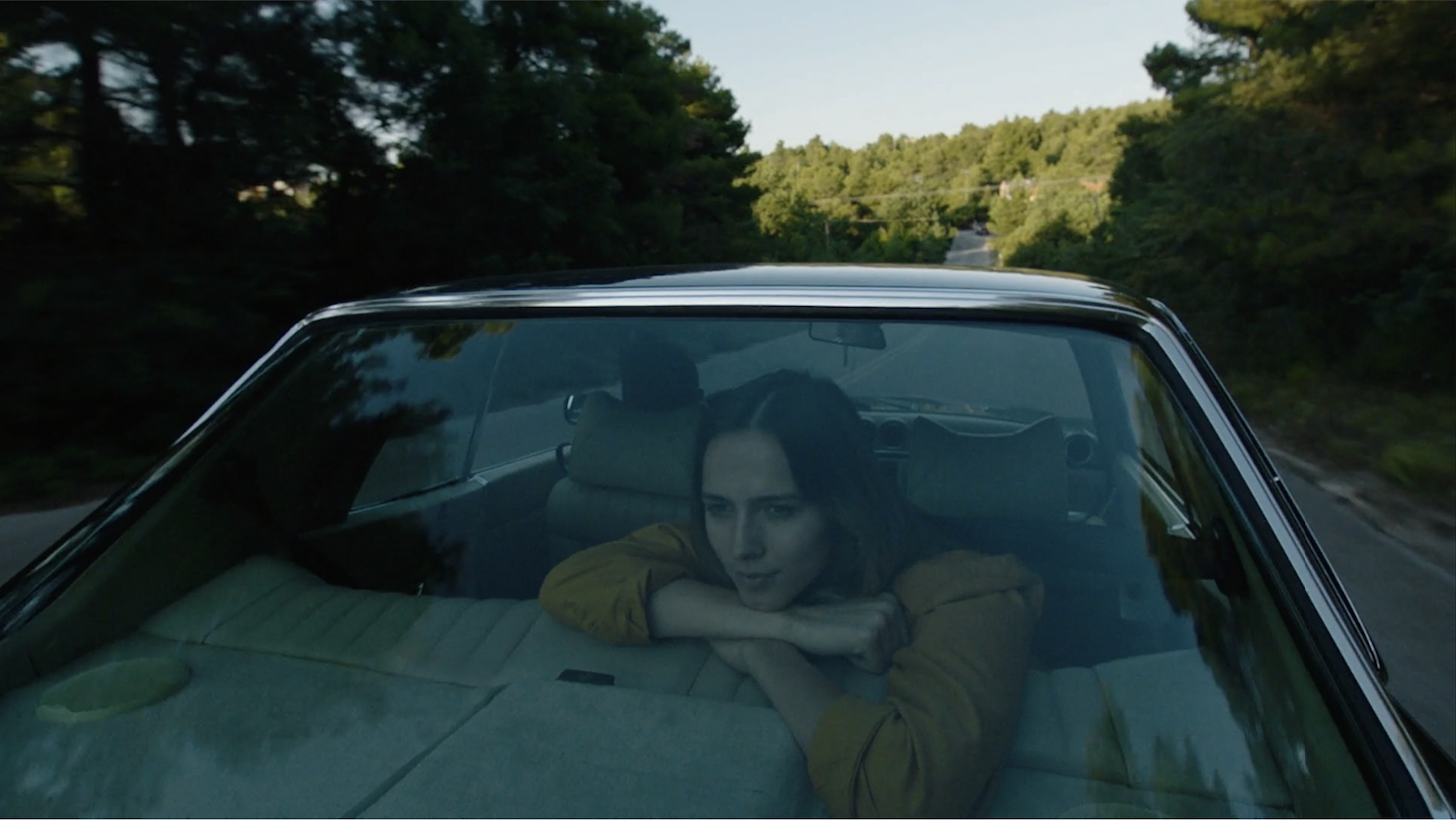 Woman in yellow shirt looks out the back window of a convertible driving on a rural road through a forest on a sunny day.