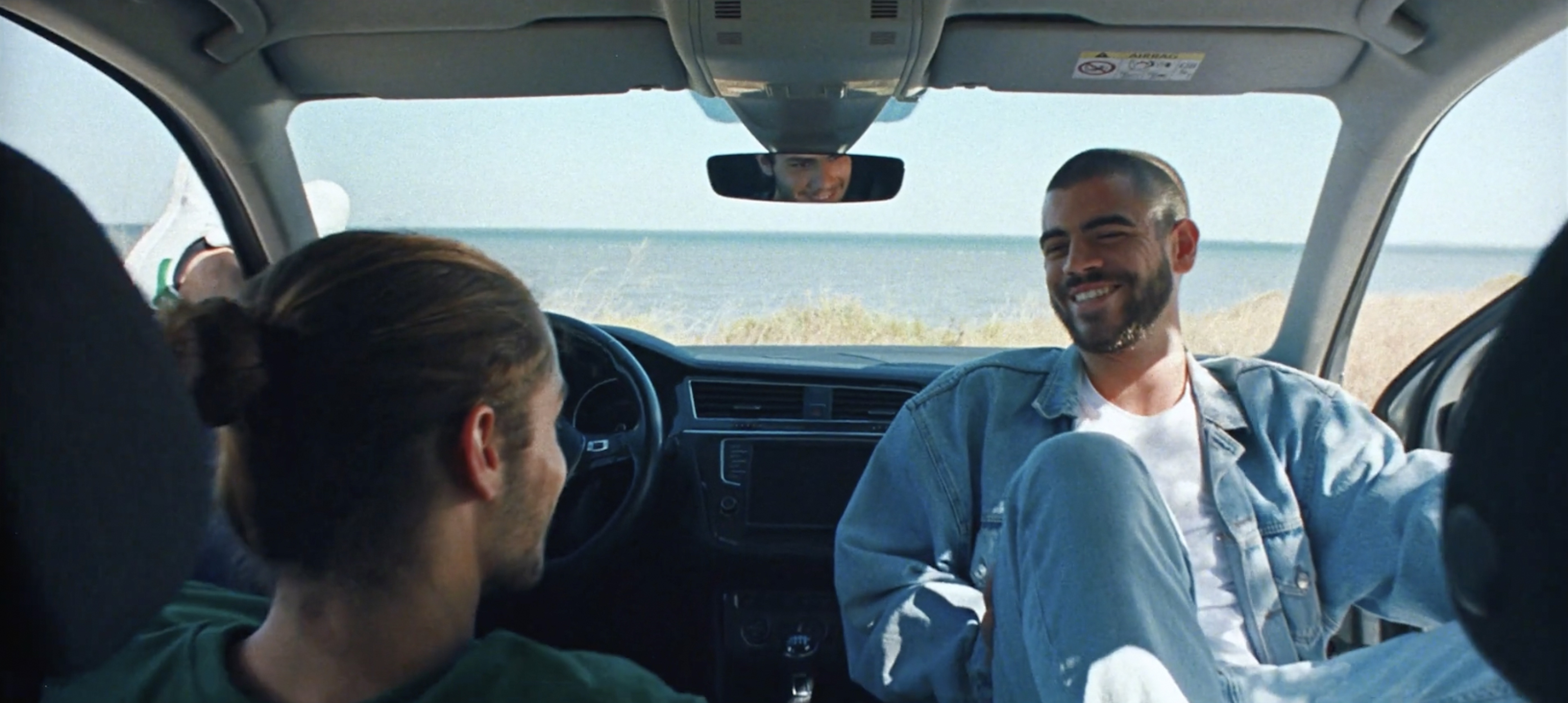 Two young adult men sit in a car with a view of the ocean and beach in the background, both smiling and wearing casual clothing.