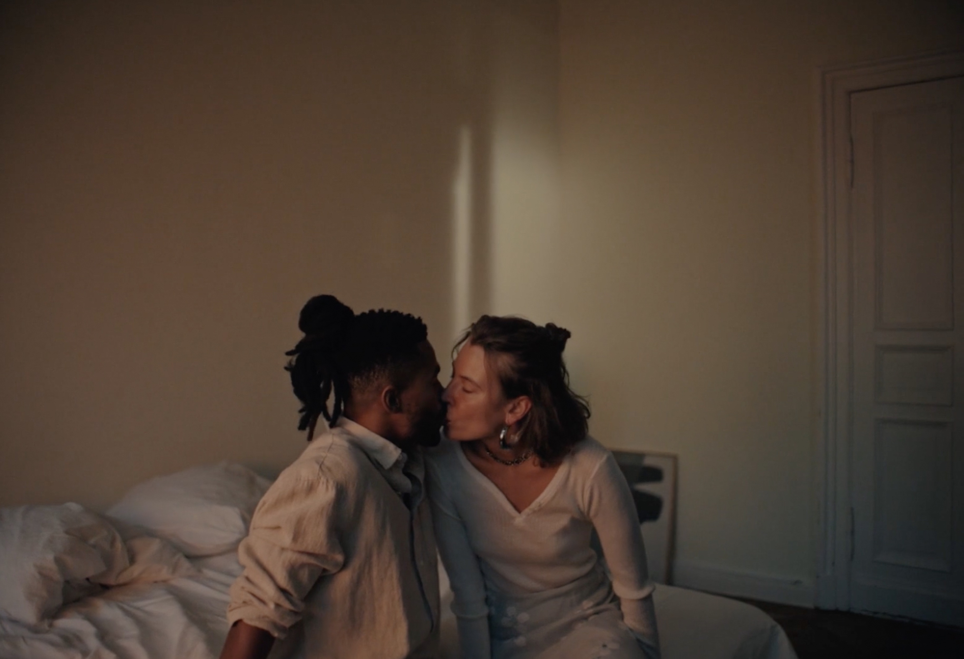 A romantic moment captured indoors: an adult man and woman in white, sharing a kiss on a bed with neutral walls and soft, diffused lighting.