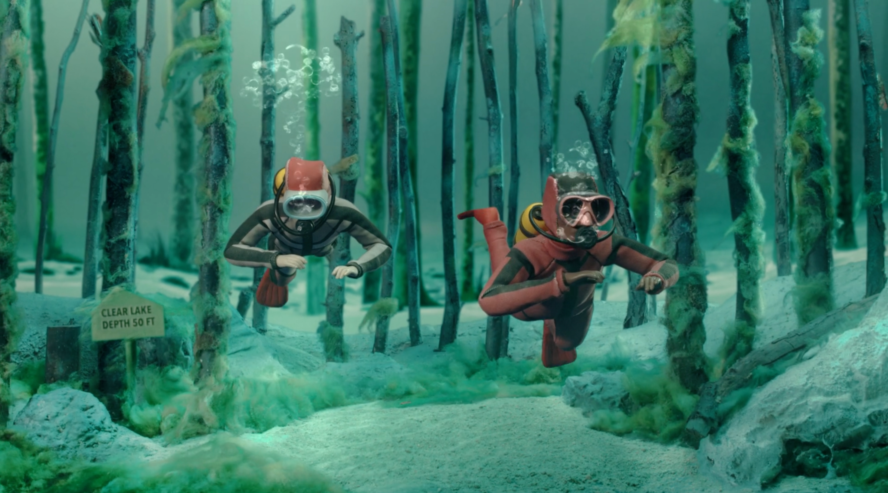 Two scuba divers swim underwater among tree trunks in a lake, with air bubbles rising and a sign indicating the lake's depth.