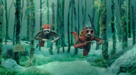 Two divers in red and dark wetsuits swimming through an underwater forest diorama with a wooden depth sign