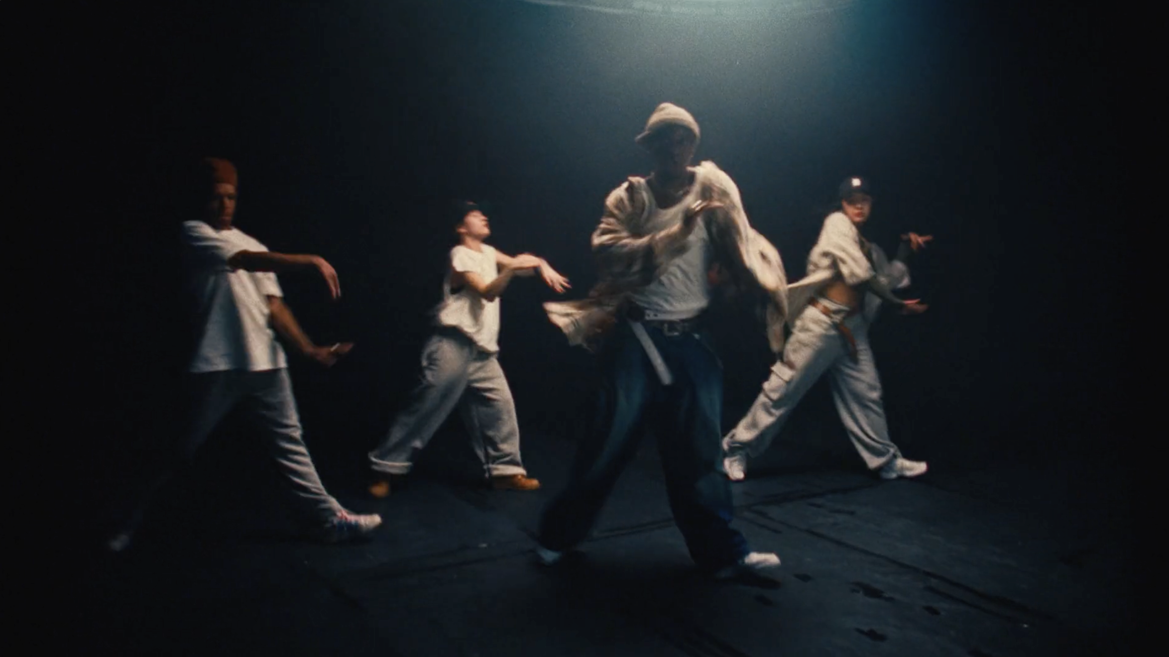 A diverse group of four dancers in relaxed street wear performing a choreographed dance routine in a dimly lit studio space with dramatic lighting.
