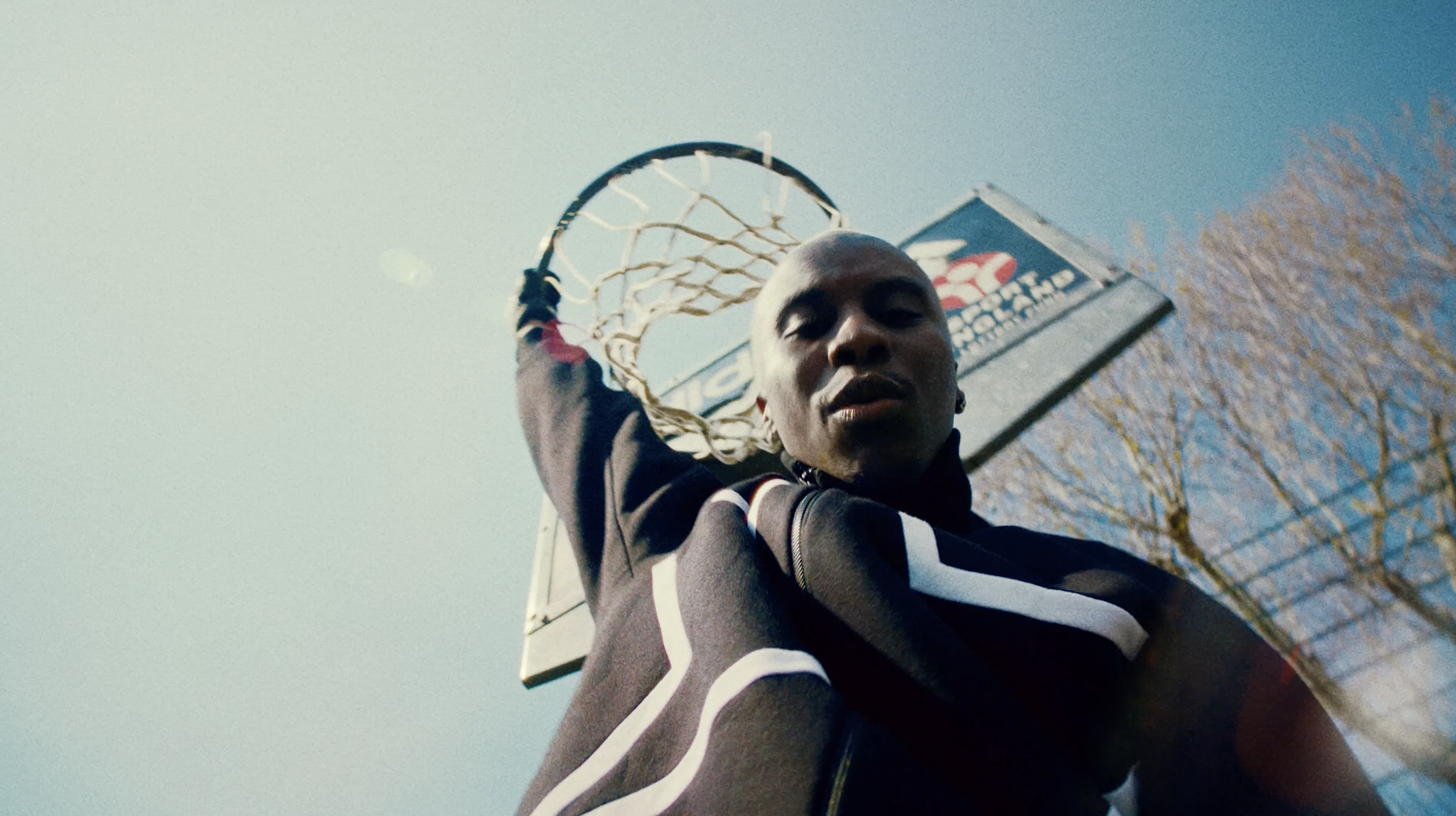 Adult male basketball player with a dark complexion wears a black and white jacket, standing outdoors under a basketball hoop and clear sky.