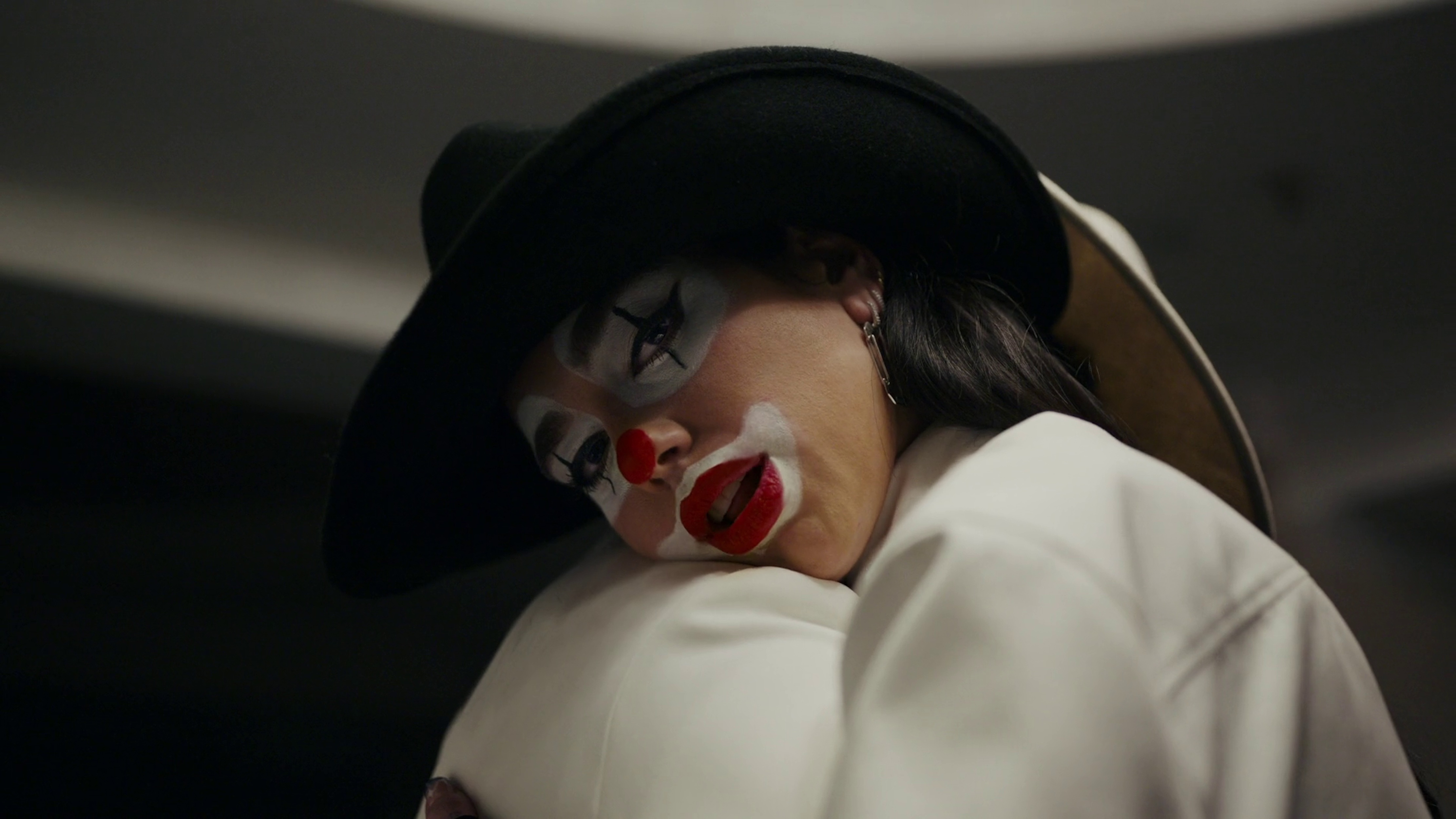 A woman with clown makeup and a black hat is embracing someone while indoors. Her face has white foundation and she has red lipstick.