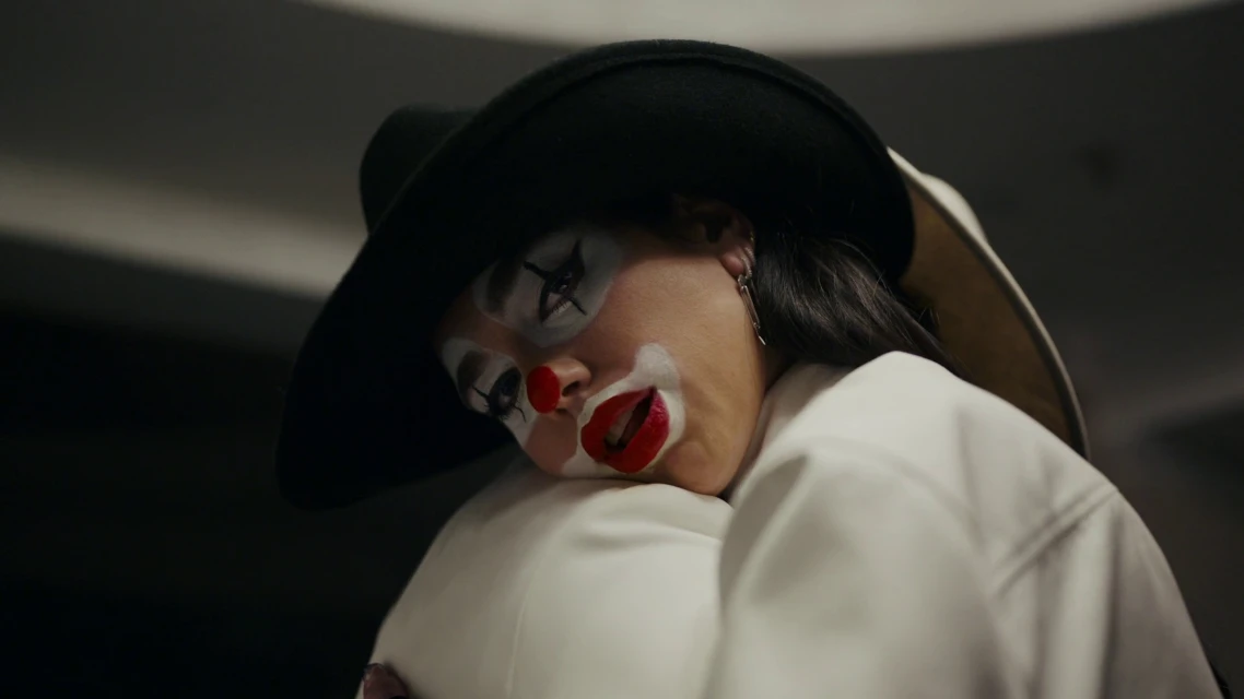 A woman with clown makeup and a black hat is embracing someone while indoors. Her face has white foundation and she has red lipstick.