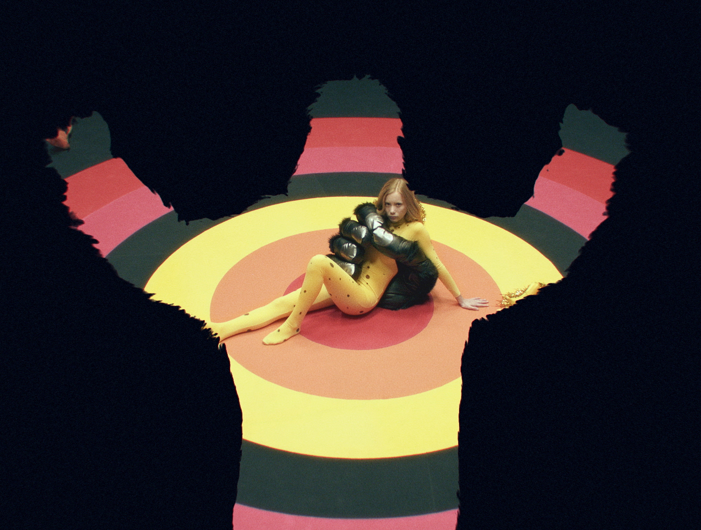A woman in a yellow spotted bodysuit reclines in front of a large black gorilla hand inside a target graphic with concentric bands.