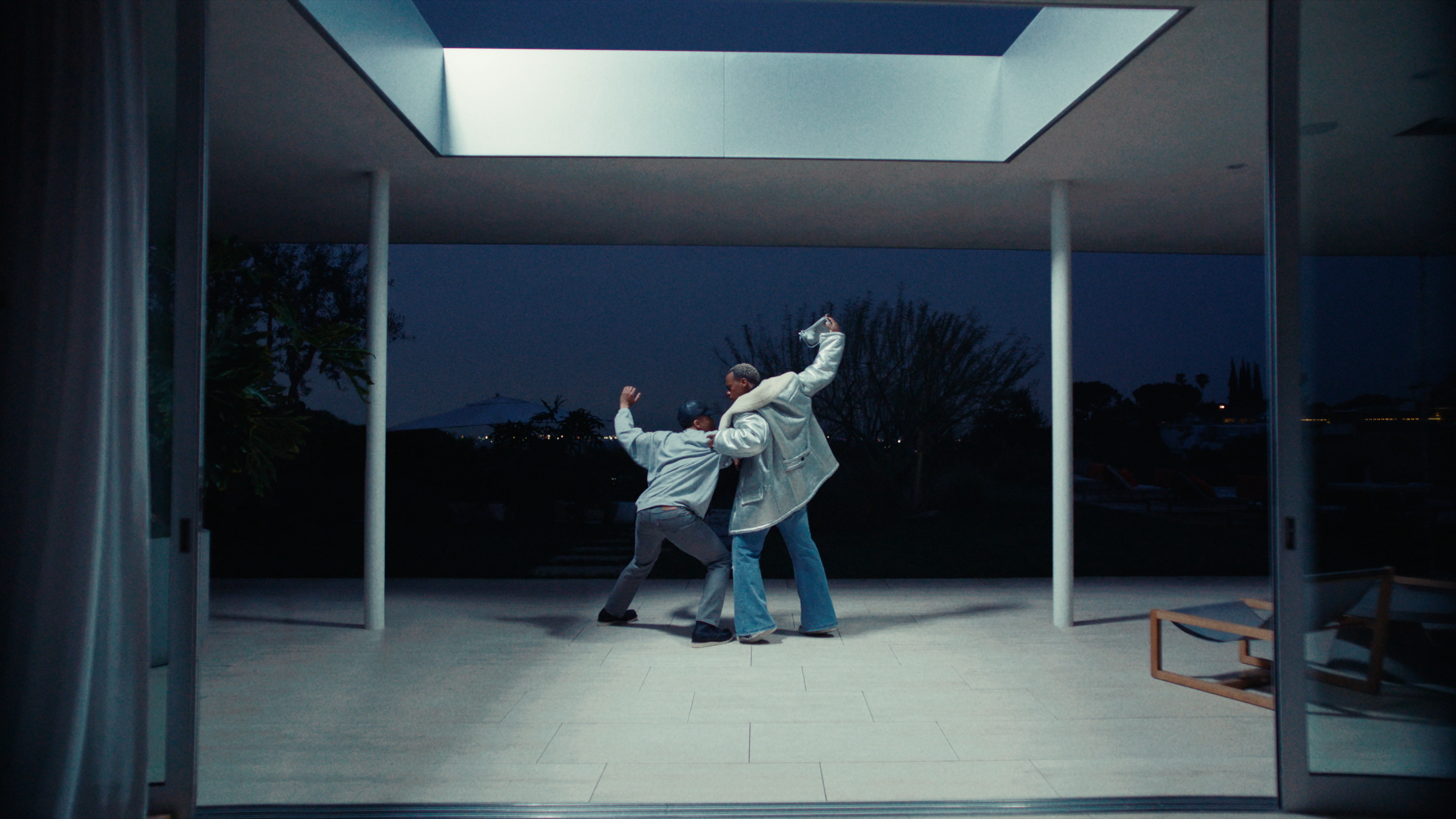 Two adult males in a modern house with a courtyard dance during the night, with an exterior view through the open walls and skylight above.