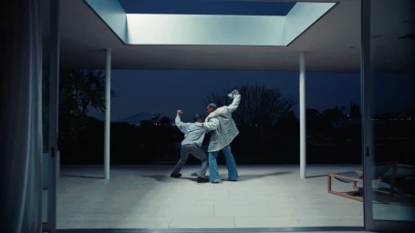 Two men dancing inside a modern minimalist home at night viewed through a wide glass sliding door opening