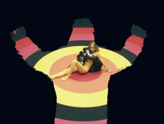 Woman in yellow catsuit seated on a target-patterned floor framed by large dark silhouette shapes directed by Lope Serrano