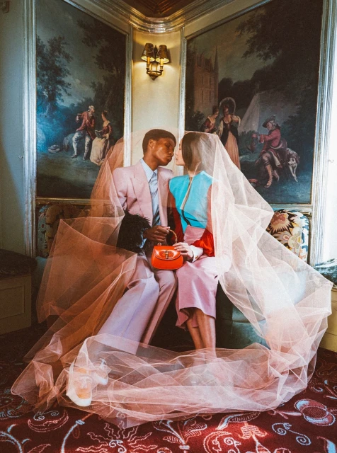A couple poses indoors with a veil. The pair are in retro suits sitting on furniture in front of mural art in muted lighting.