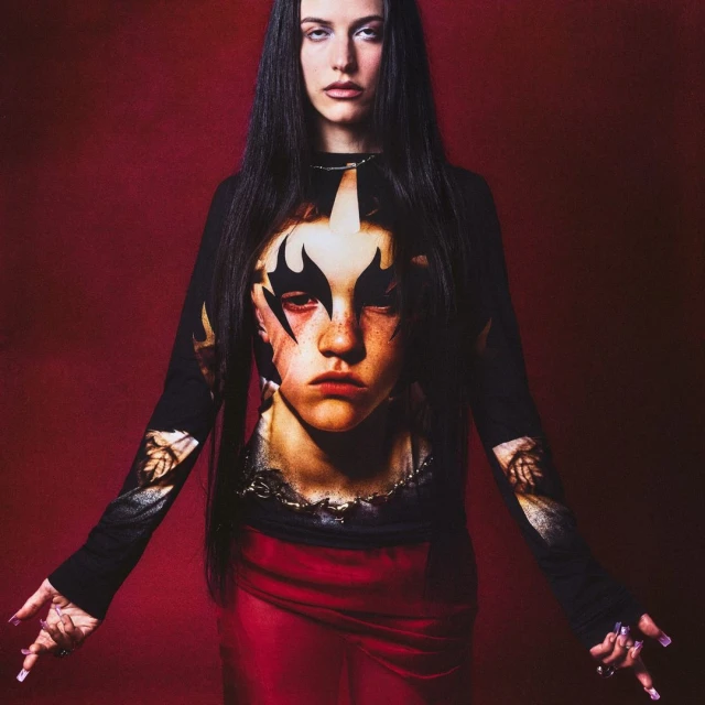 Portrait of a woman with long black hair wearing a long-sleeved shirt with a face print and a long red skirt against a red backdrop.
